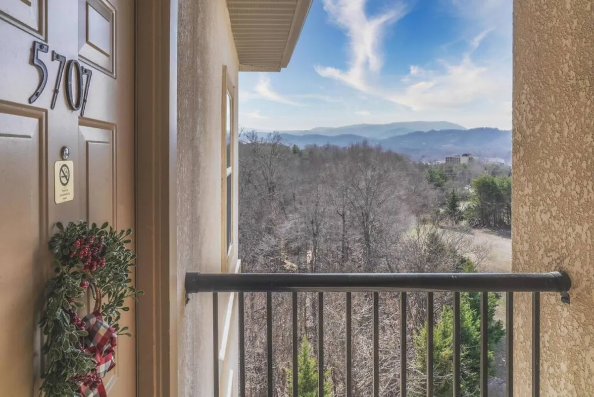 The image shows a welcoming front door adorned with a wreath, and beyond the balcony railing, expansive mountain views stretch into the distance. The scene is bathed in natural light, emphasizing the connection between the private space and its inspiring surroundings.