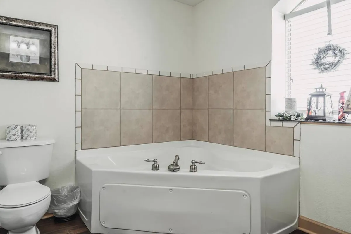 A spacious bathroom with a jacuzzi tub, framed artwork, and a window adorned with decorative items. The atmosphere is tranquil, offering a luxurious spot to unwind and indulge in personal downtime.