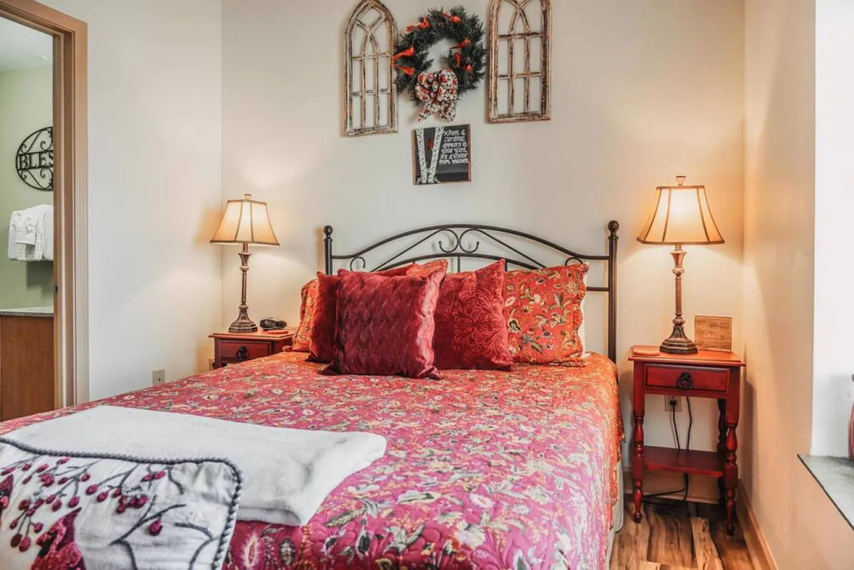 A warmly lit bedroom featuring a bed with patterned bedding, decorative pillows, table lamps, and a wreath on the wall. The ambiance is inviting and perfect for unwinding after a busy day. The room is suited for adults or couples, showcasing a sense of peaceful retreat.