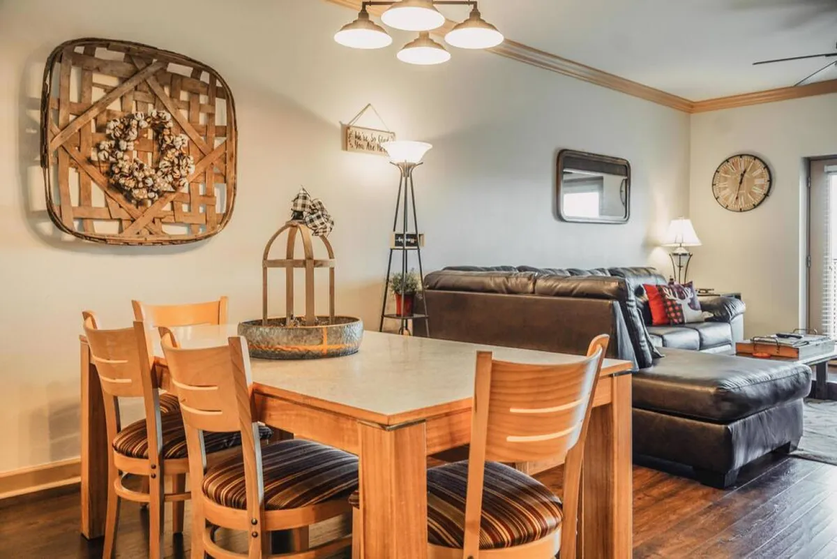 A cozy dining table sits adjacent to a sofa, surrounded by decorative wall art, a floor lamp, and a classic clock. The ambiance is intimate and welcoming, ideal for family dinners or game nights, all within sight of the living space.