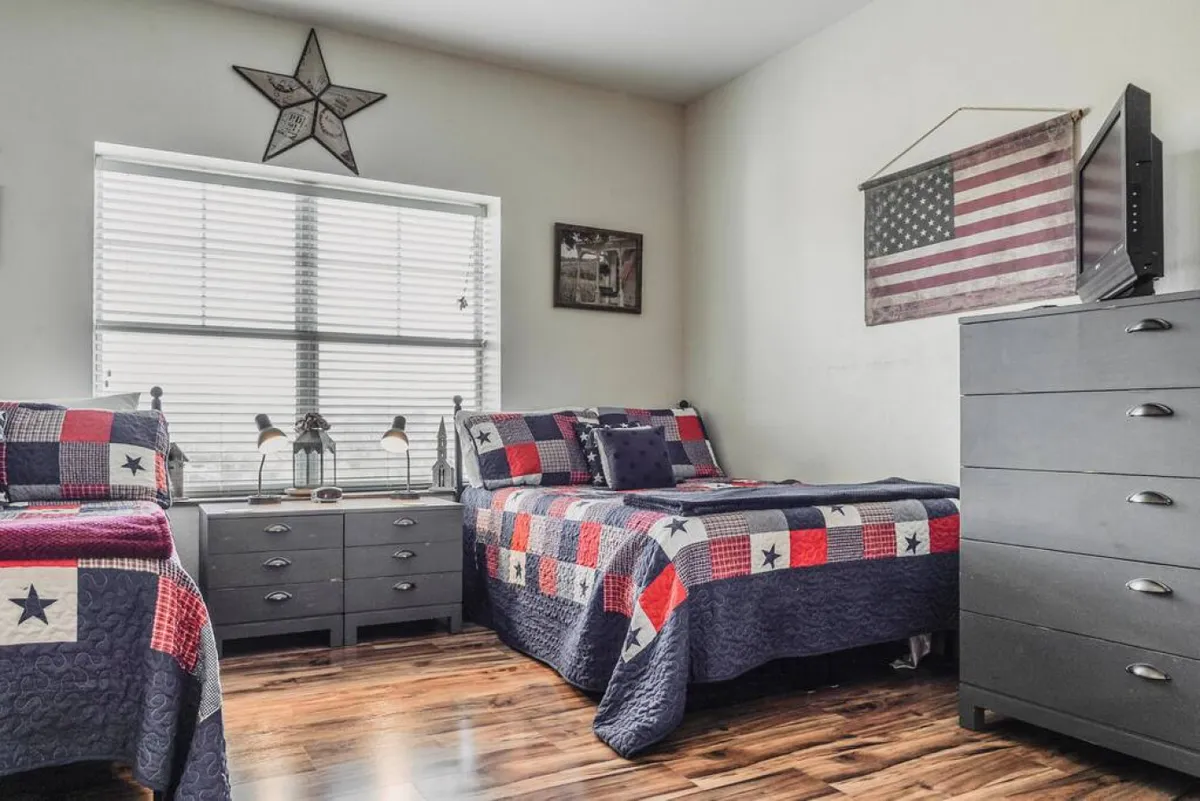 A cheerful twin bedroom with two beds featuring colorful quilts, star-themed wall decor, and a dresser topped with a television. Sunlight filters through the window, adding a bright, playful feel ideal for younger guests.