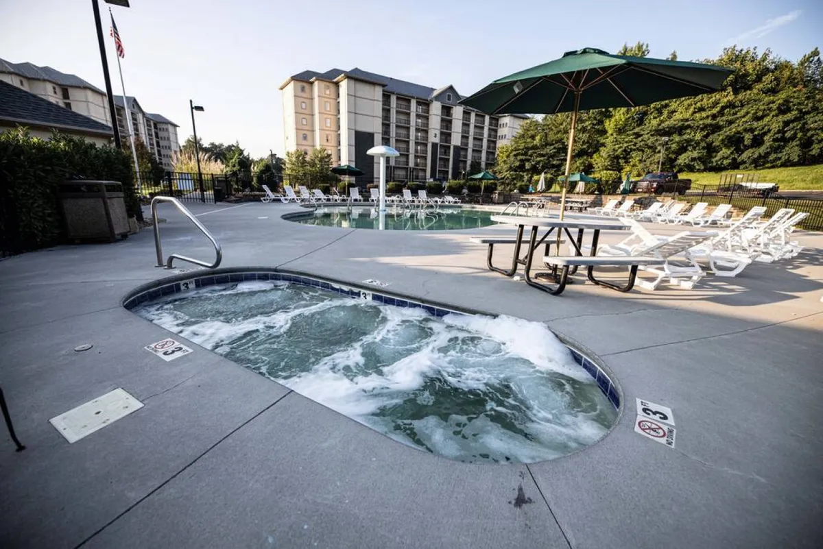 The image features an exterior view of the pool and hot tub area, with a picnic table shaded by a green umbrella and plenty of lounge chairs. The setting exudes a leisurely, social vibe, ideal for relaxing after a day of activities or enjoying a family picnic.