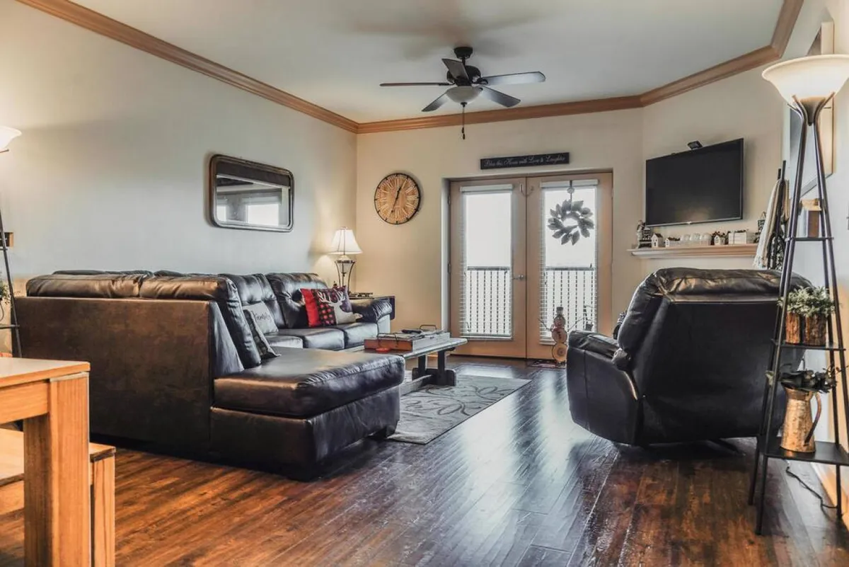 A cozy living area with a leather sofa and coffee table, illuminated by natural light streaming through balcony doors. The decorative clock and soft lighting create an inviting, home-like ambiance, while the view through the doors hints at outdoor spaces and the surrounding scenery.